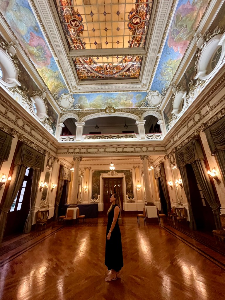 Interior de um salão histórico com arquitetura neoclássica, teto ornamentado com pinturas e vitrais, piso de madeira e Dra. Wyara contemplando o ambiente.